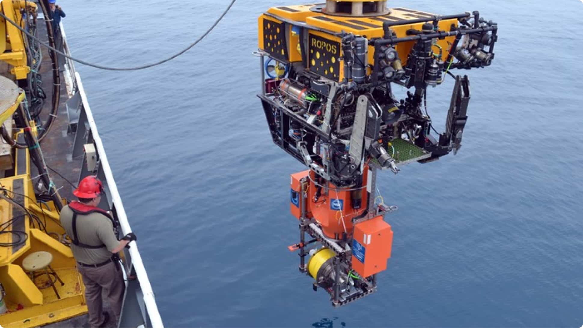 An underwater Remote Operated Vehicle (ROV) is lowered into the ocean from a large research vessel as a person in a hard hat watches, a large profiler instrument is attached to the bottom of the ROV