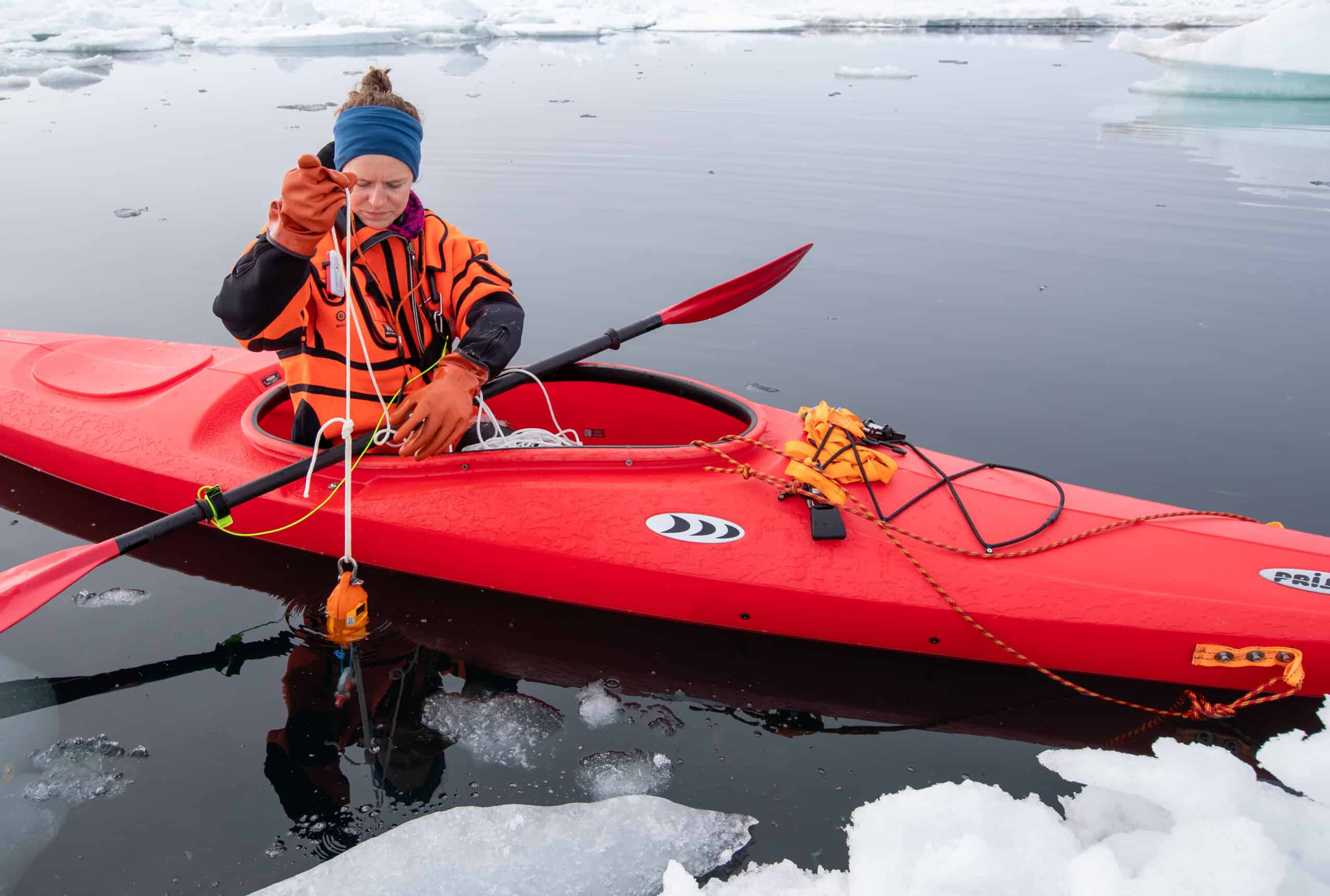 A researcher is sitting in a kayak holding a line attached to a scientific instrument, the water is calm, with large pieces of ice in the foreground and background
