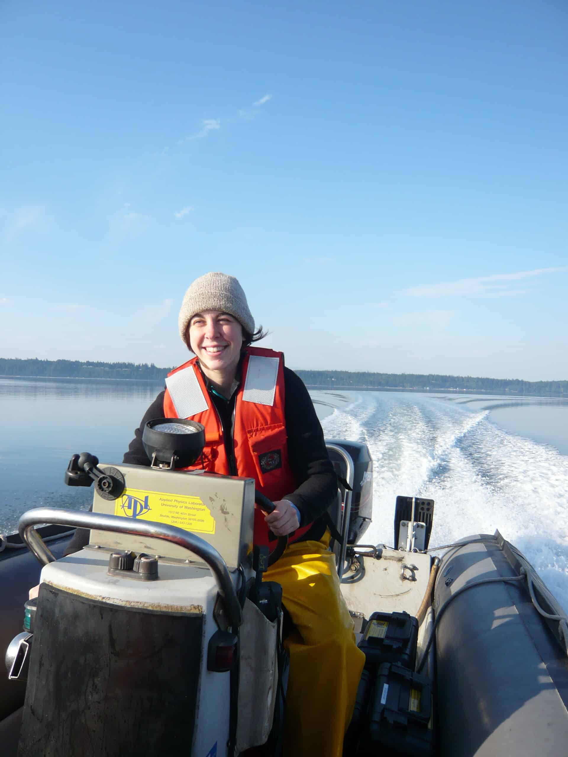 A researcher is piloting an inflatable powered boat, smiling and facing into the sun, with the boat's wake and shoreline in the background