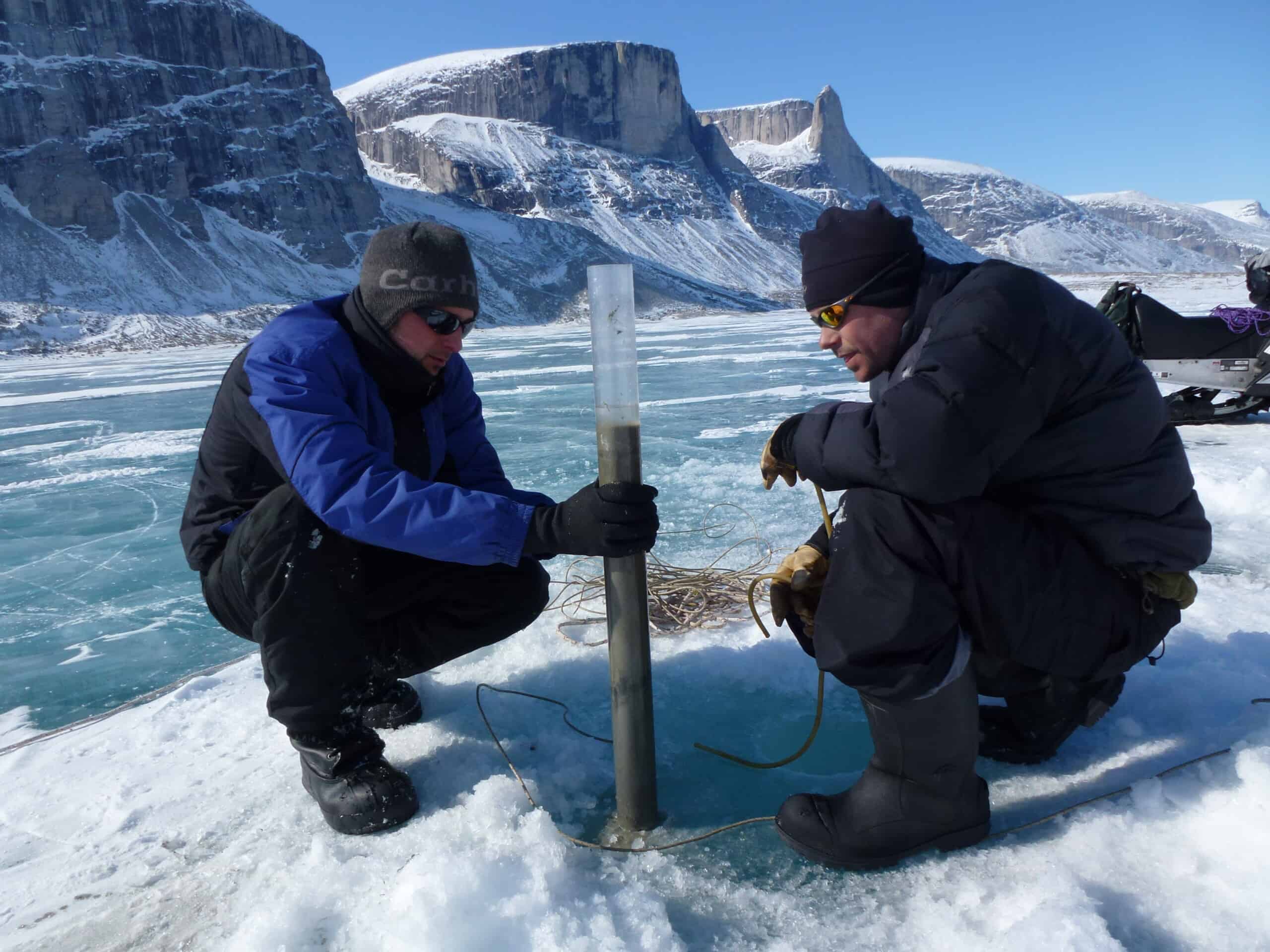 Two men in snow suits and sunglasses are crouched on ice taking an ice core sample, snow-covered mountain cliffs in background.