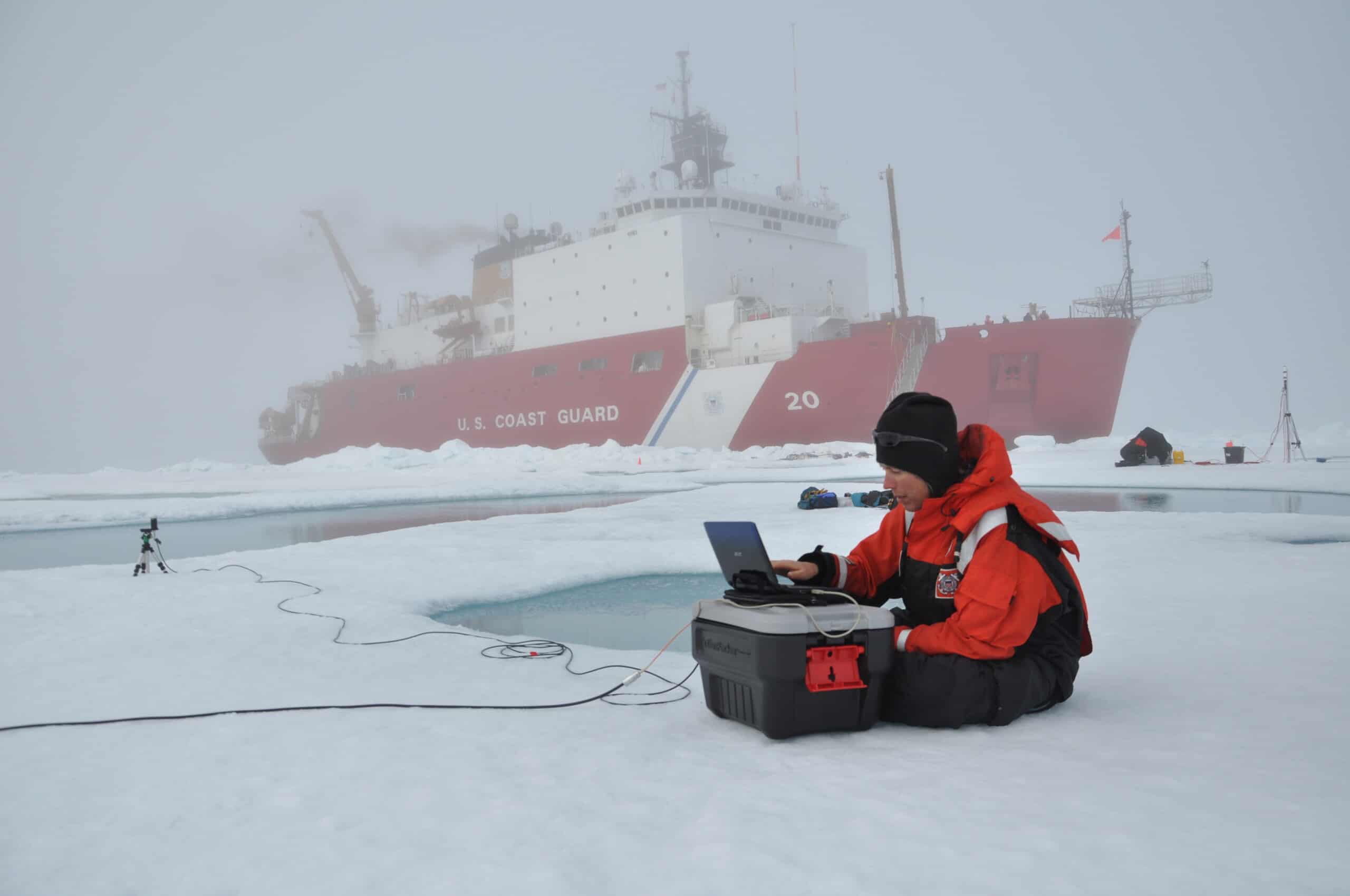 A researcher wearing an ice suit sits on the ice viewing a laptop that is connected to an instrument by a cable, the US Coast Guard Cutter Healy in the background