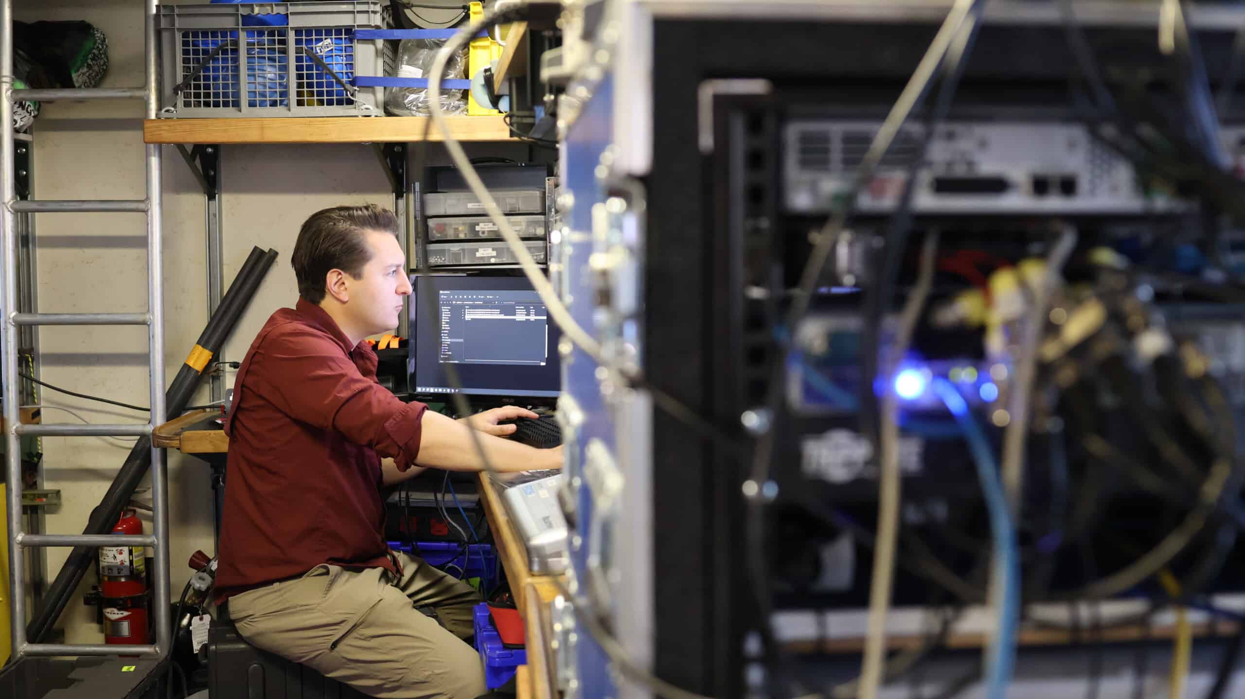 Computer equipment and cables are in foreground, a person viewing a computer screen in the background